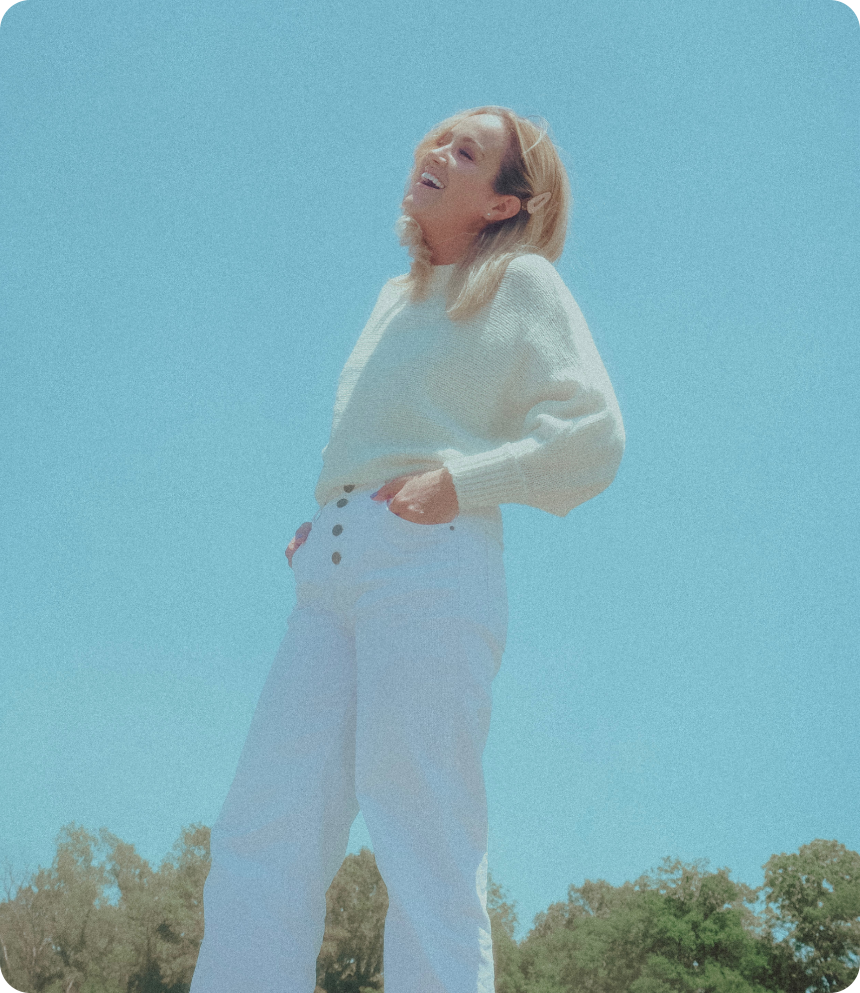 A woman in a white sweater and white pants is standing in front of a blue sky.