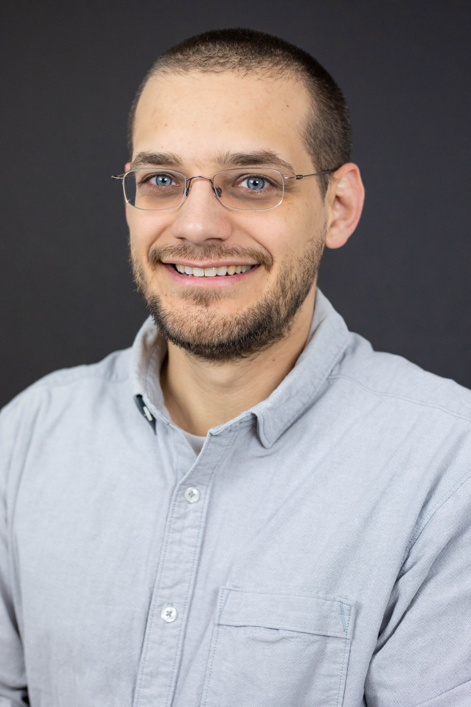 A man wearing glasses and a grey shirt is smiling for the camera.
