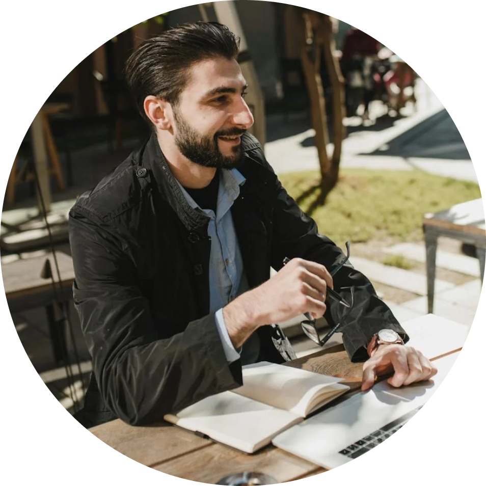 A man with a beard is sitting at a table with a laptop and a notebook.