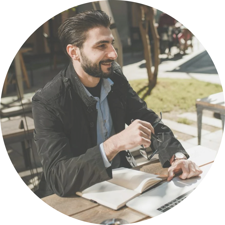 A man with a beard is sitting at a table with a notebook and a laptop