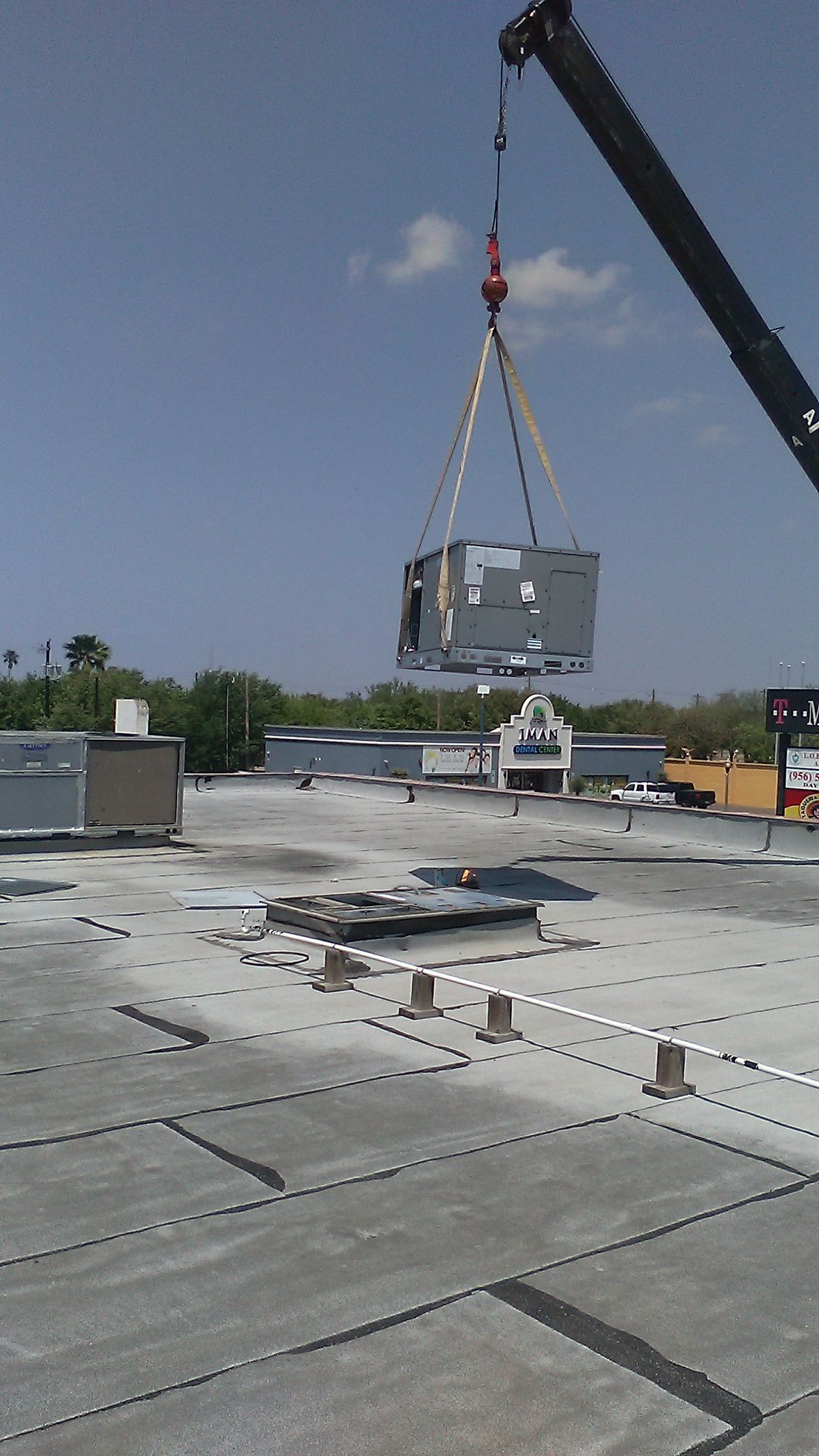 Crane lifting an HVAC unit onto a flat commercial roof, blue sky in background.