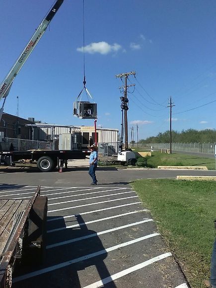 A crane lifting an HVAC unit; a worker observes. Outdoors, blue sky, parked truck.