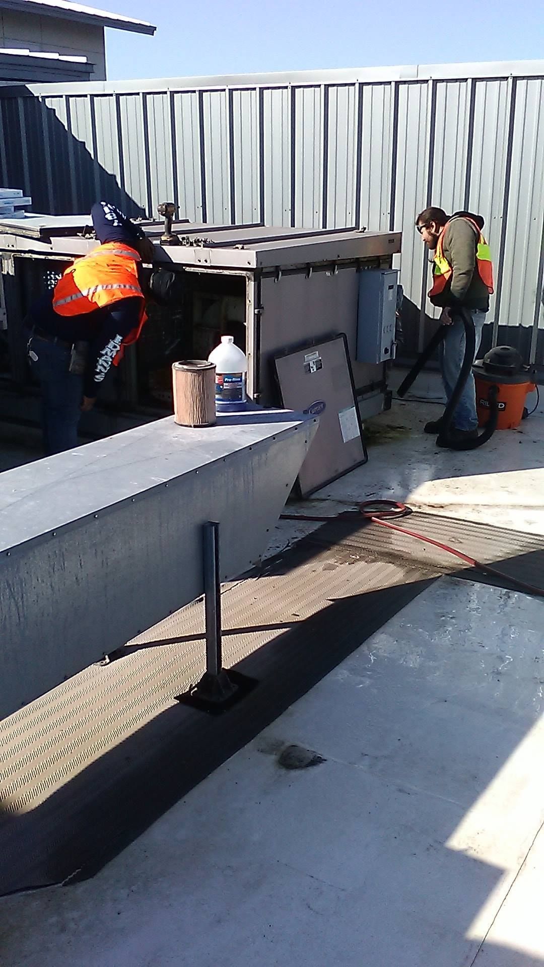 Two workers in safety vests; one grinds metal, the other uses a blower on a concrete surface outside a building.