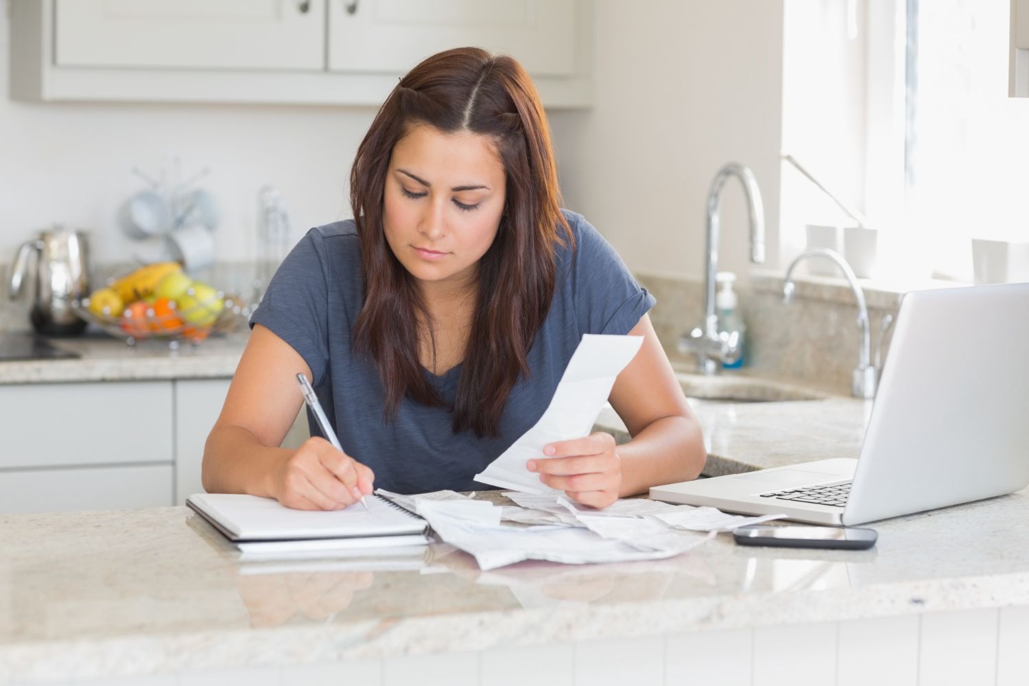 Woman in kitchen, writing on notepad, examining bills and using laptop.