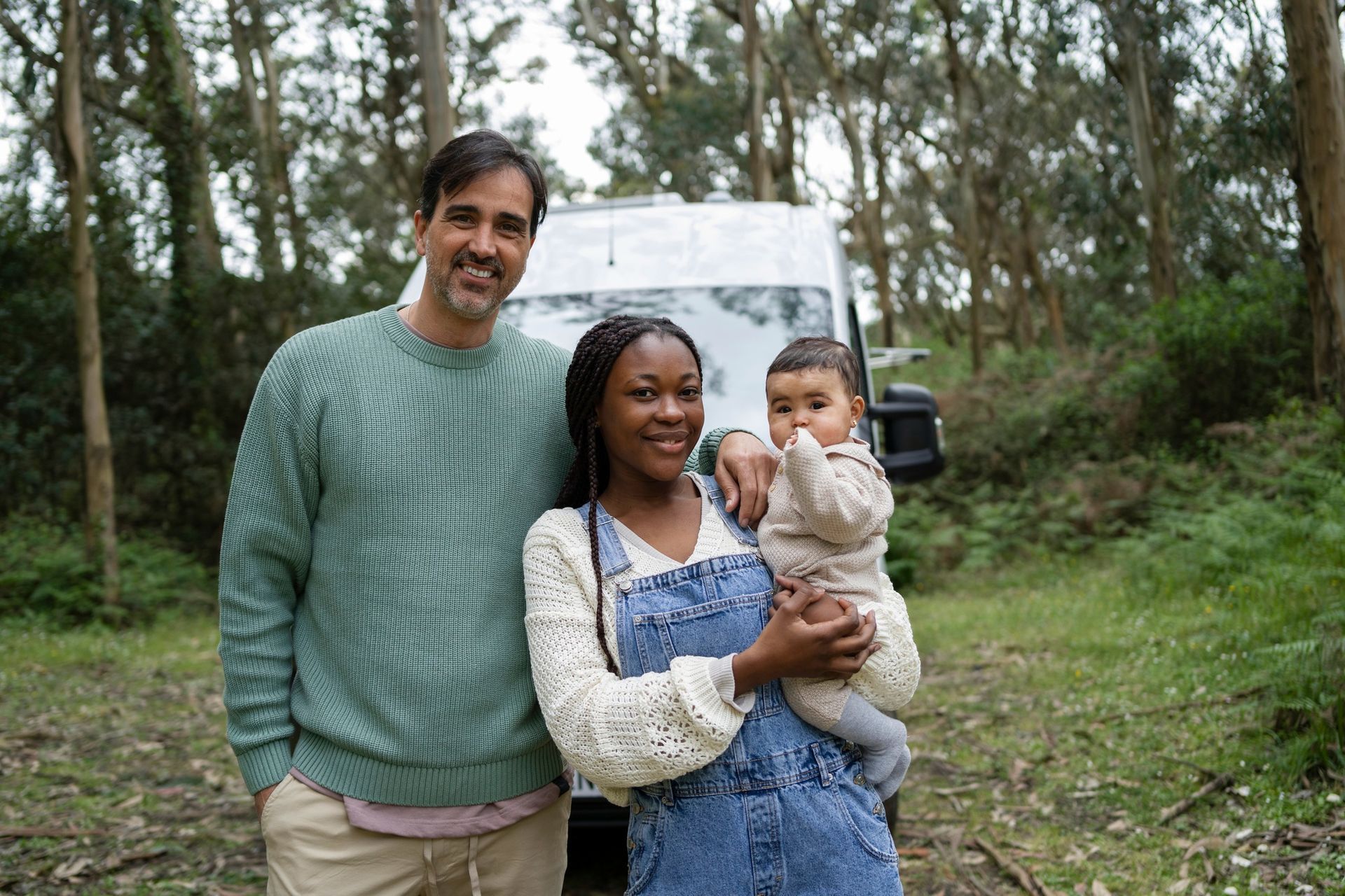 Family standing in front of a campervan in a wooded area. The woman is holding a baby.