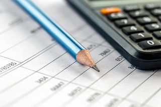 Pencil on lined paper with financial data; calculator in the background.
