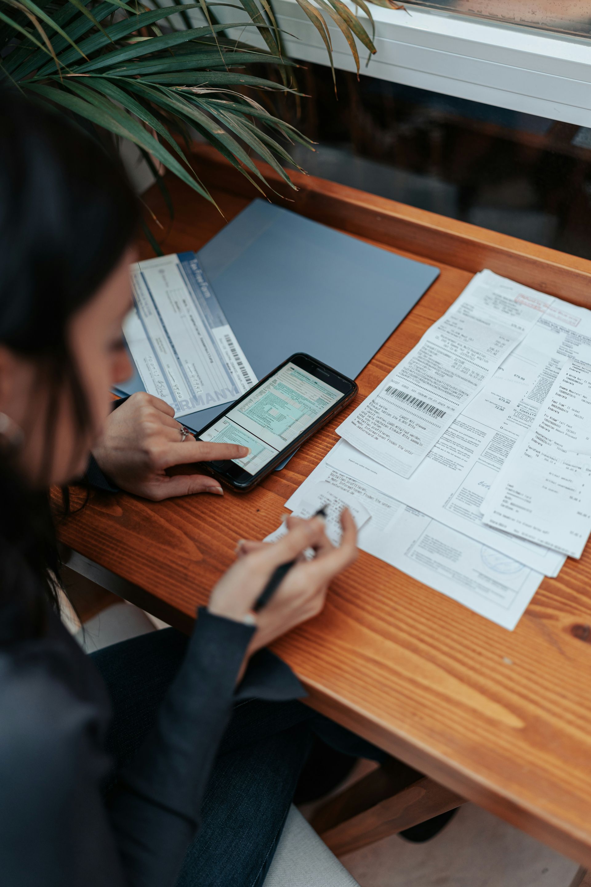 Person using a smartphone and pen, reviewing papers at a wooden desk near a window with a plant.