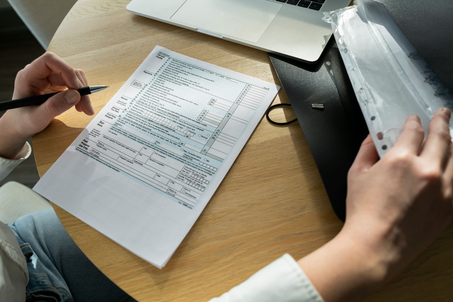 Person writing on a document at a wooden table, next to a laptop and a clear binder.