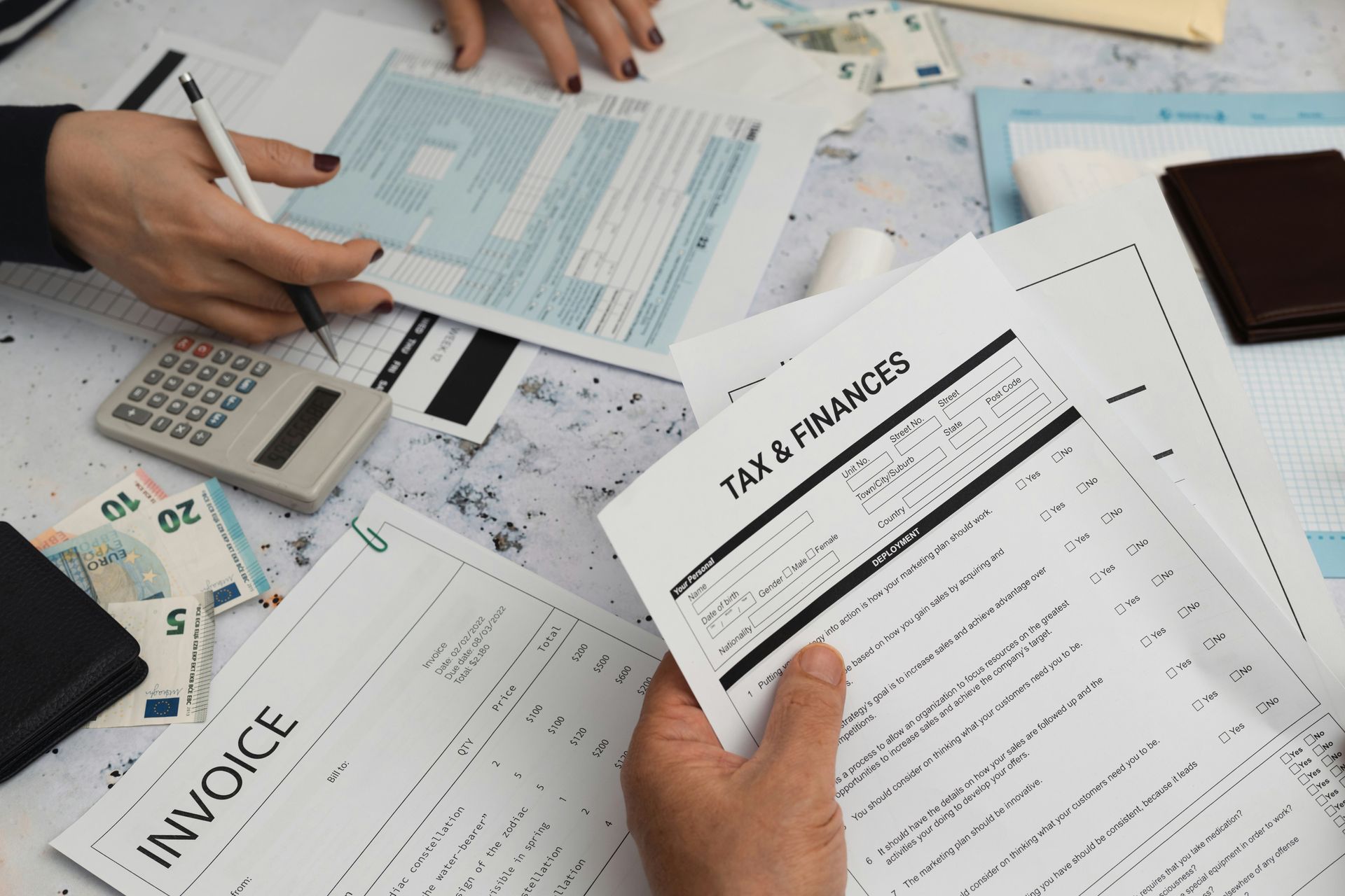 Hands reviewing tax documents, invoices, and calculator on a cluttered desk with cash and a credit card.