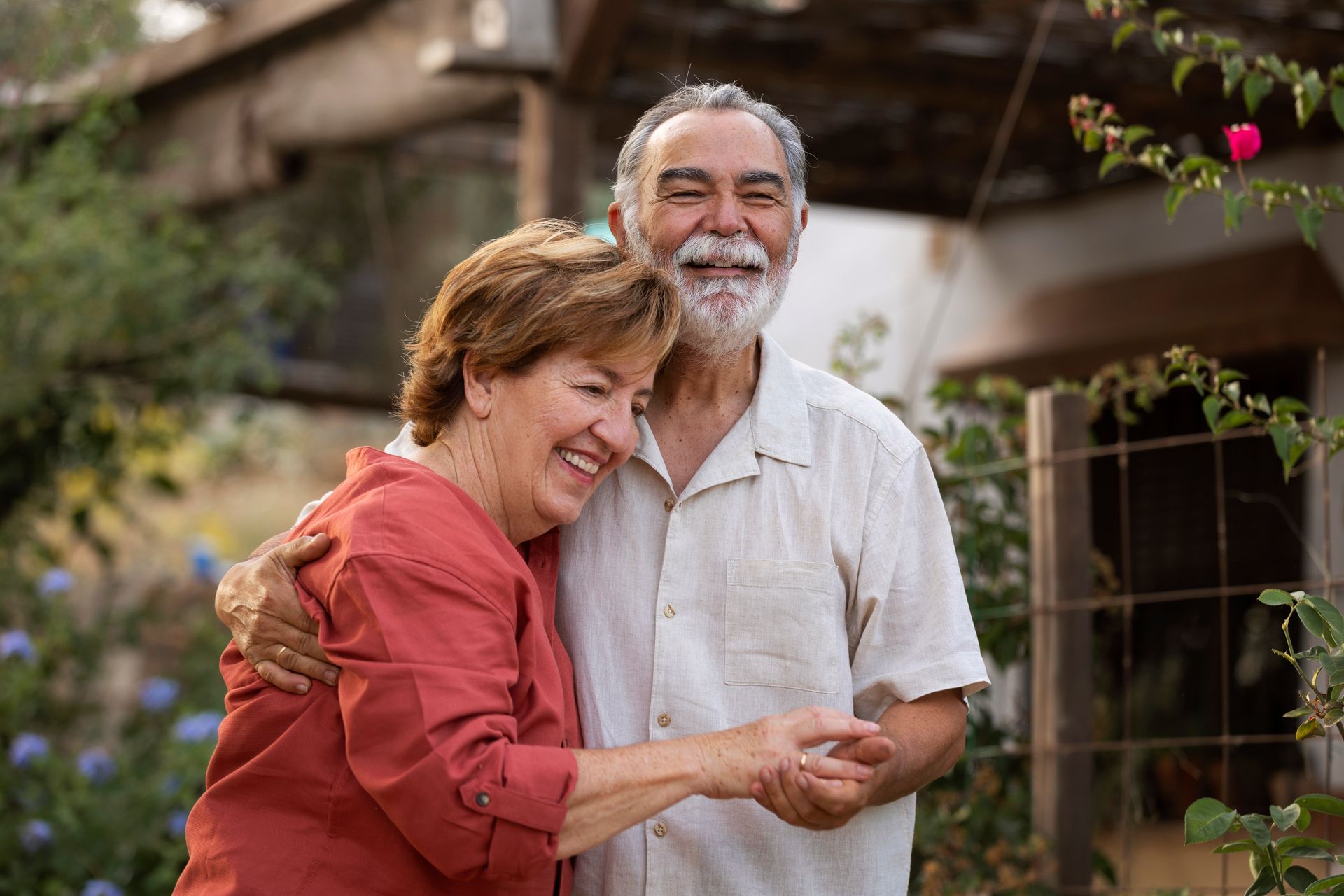 Smiling couple embracing outdoors. Woman in red shirt, man in white shirt, greenery and house in background.
