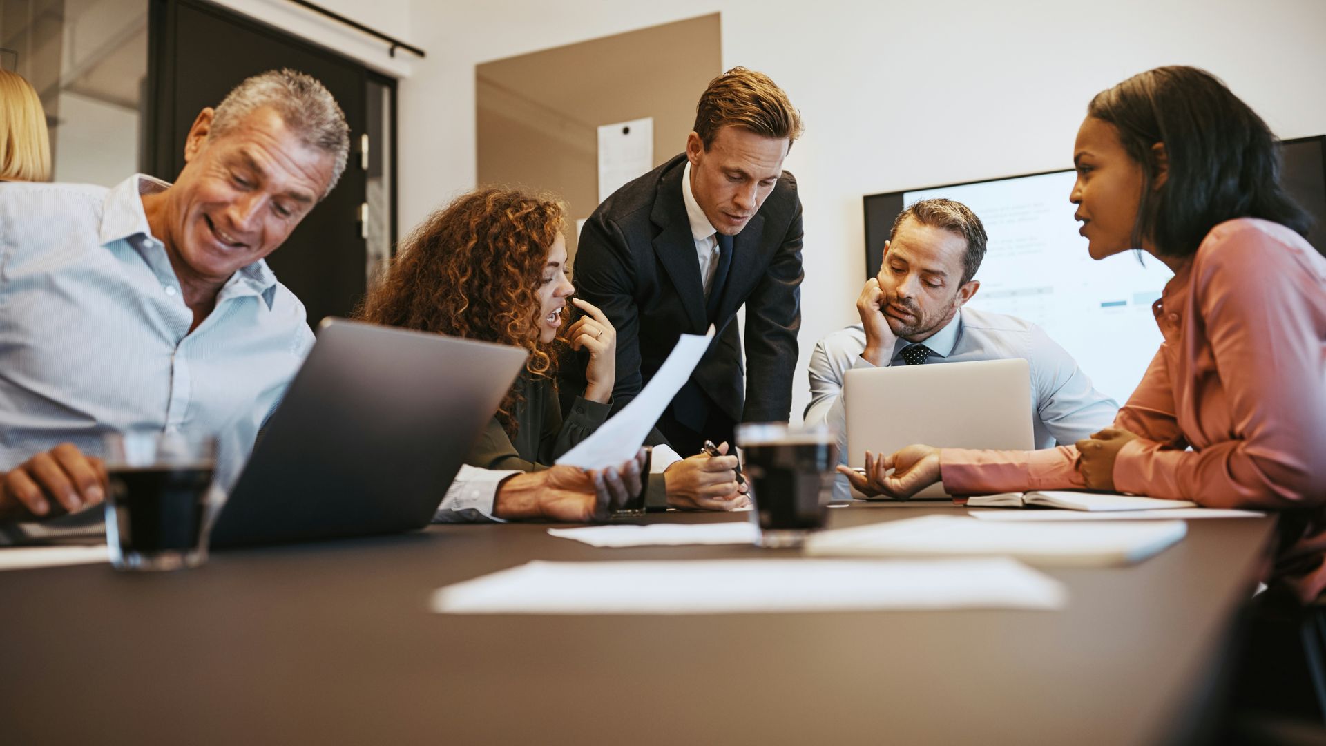 People in a meeting, reviewing documents and using laptops at a table in an office.