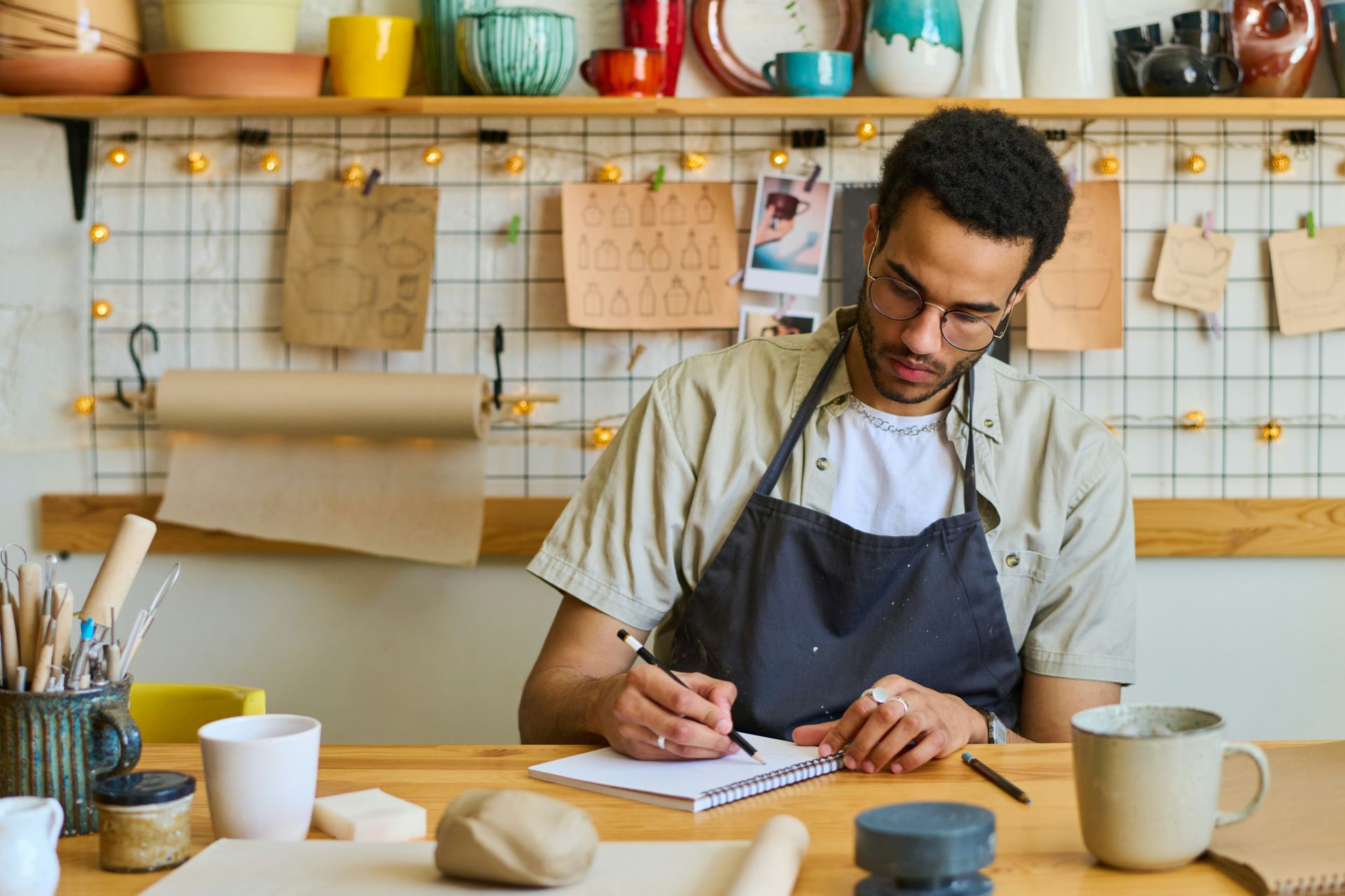 Man in apron writes in notebook at a desk, surrounded by pottery tools and materials.