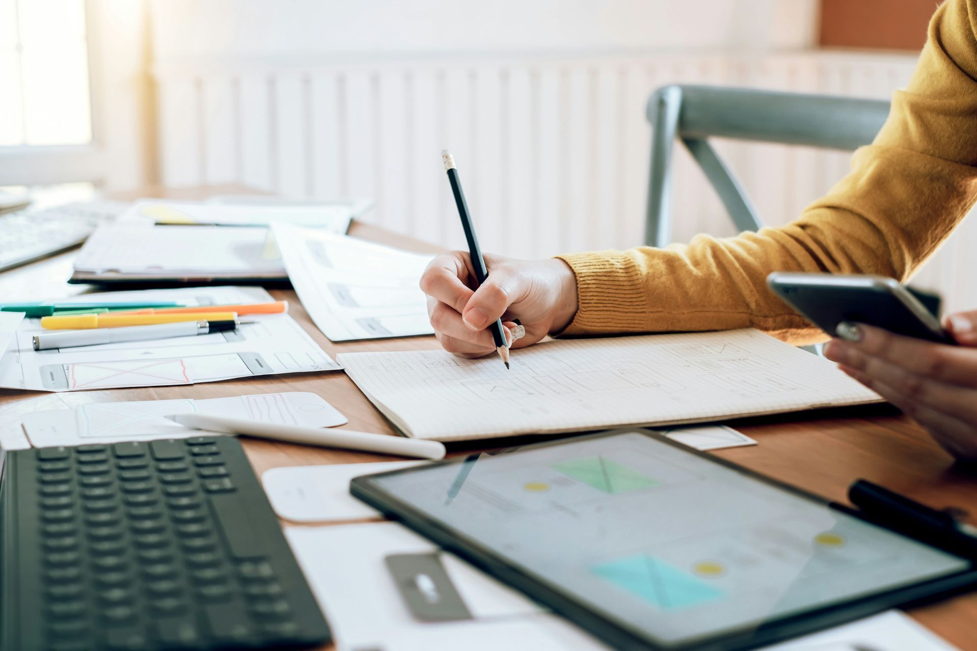 Person writing in notebook with a pencil, using a smartphone and tablet on desk.