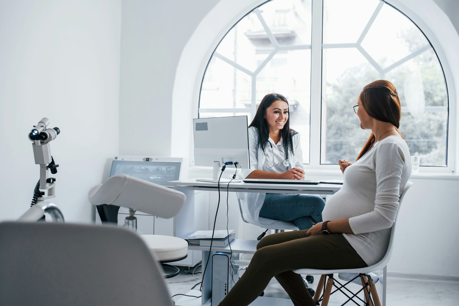 Pregnant woman at a doctor's appointment, sitting and talking with a doctor. Bright, white room with window.