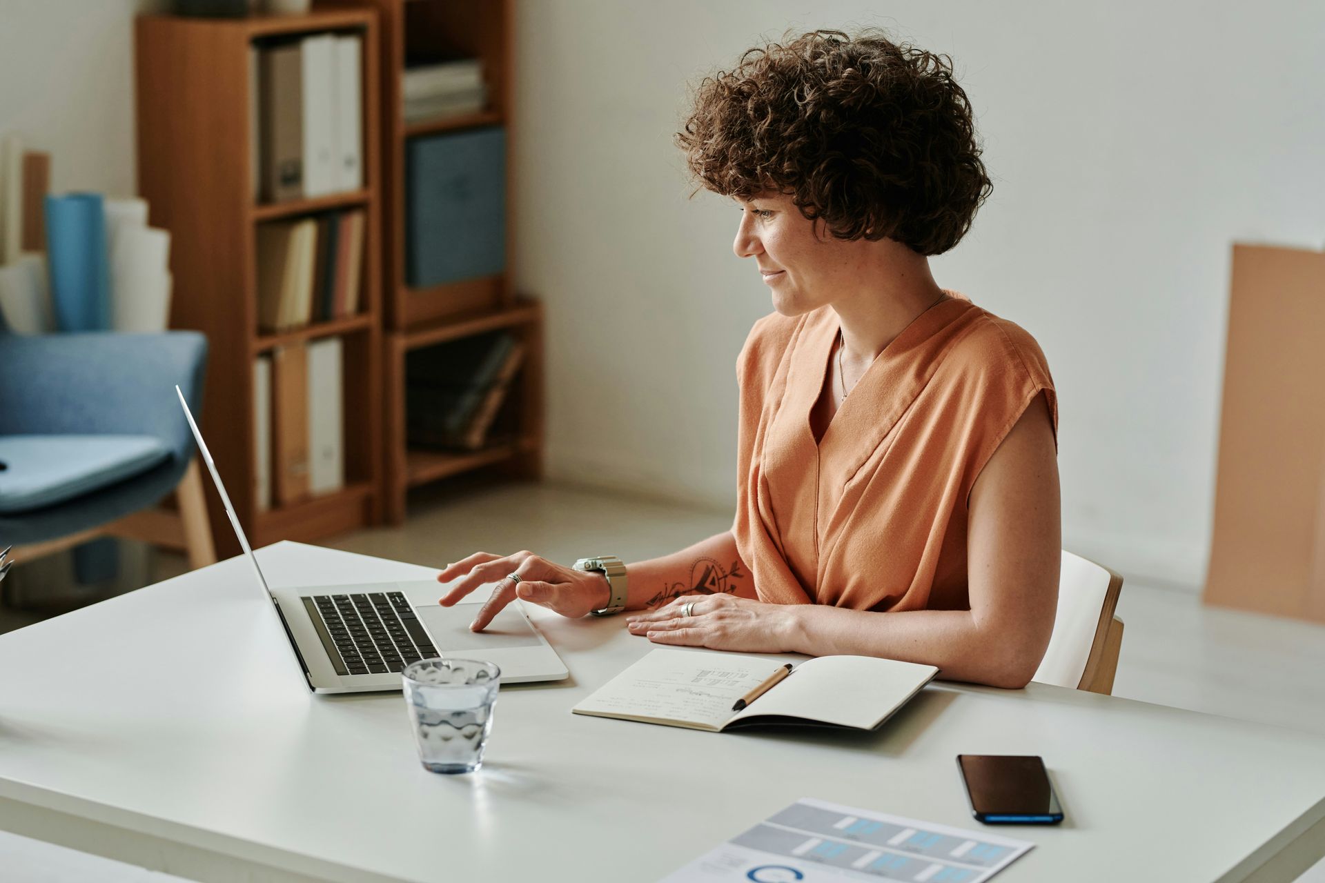 Woman with curly hair works on laptop at a desk with notebook, phone, and glass of water.
