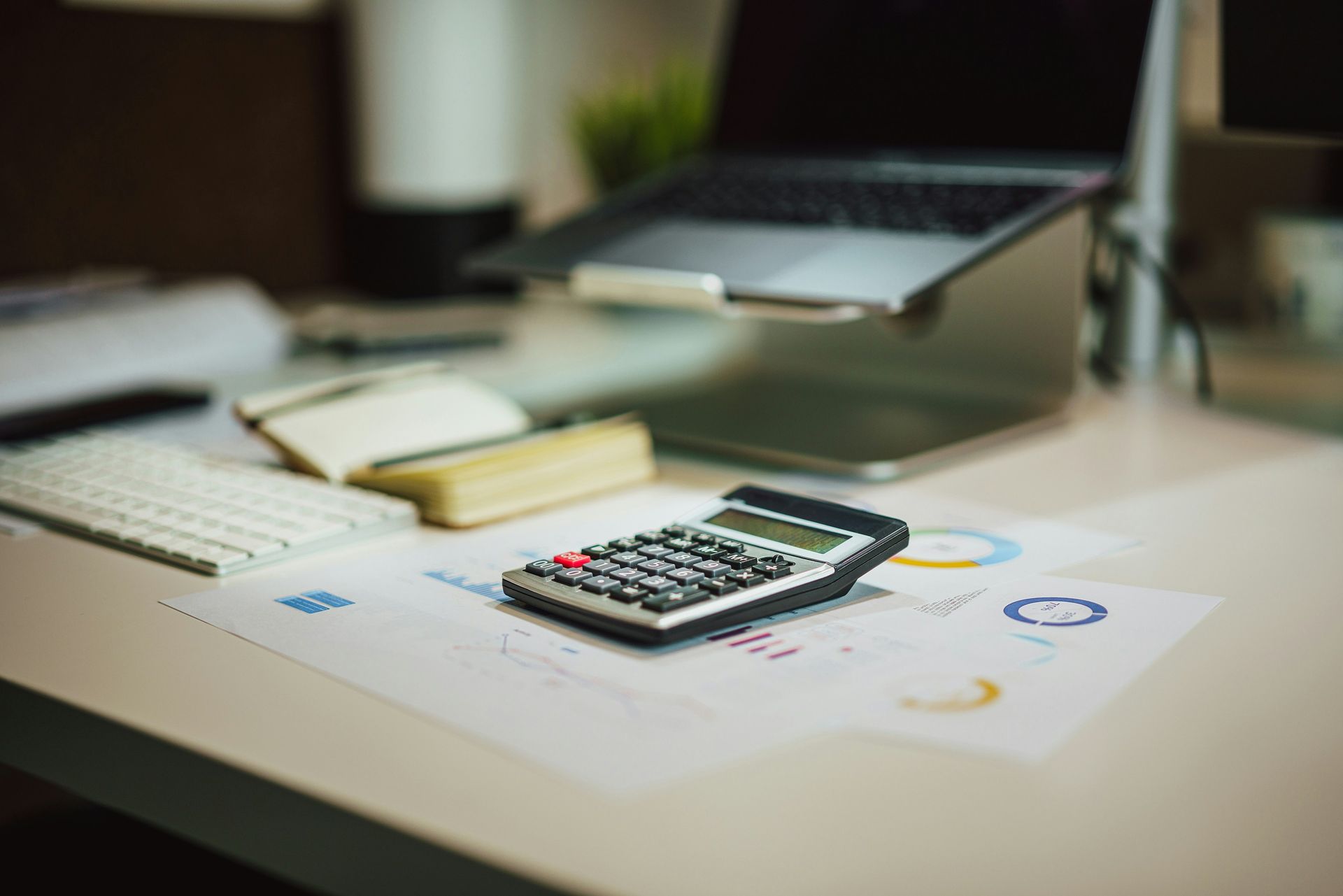 Desk with calculator, laptop on stand, keyboard, notepad, and charts.