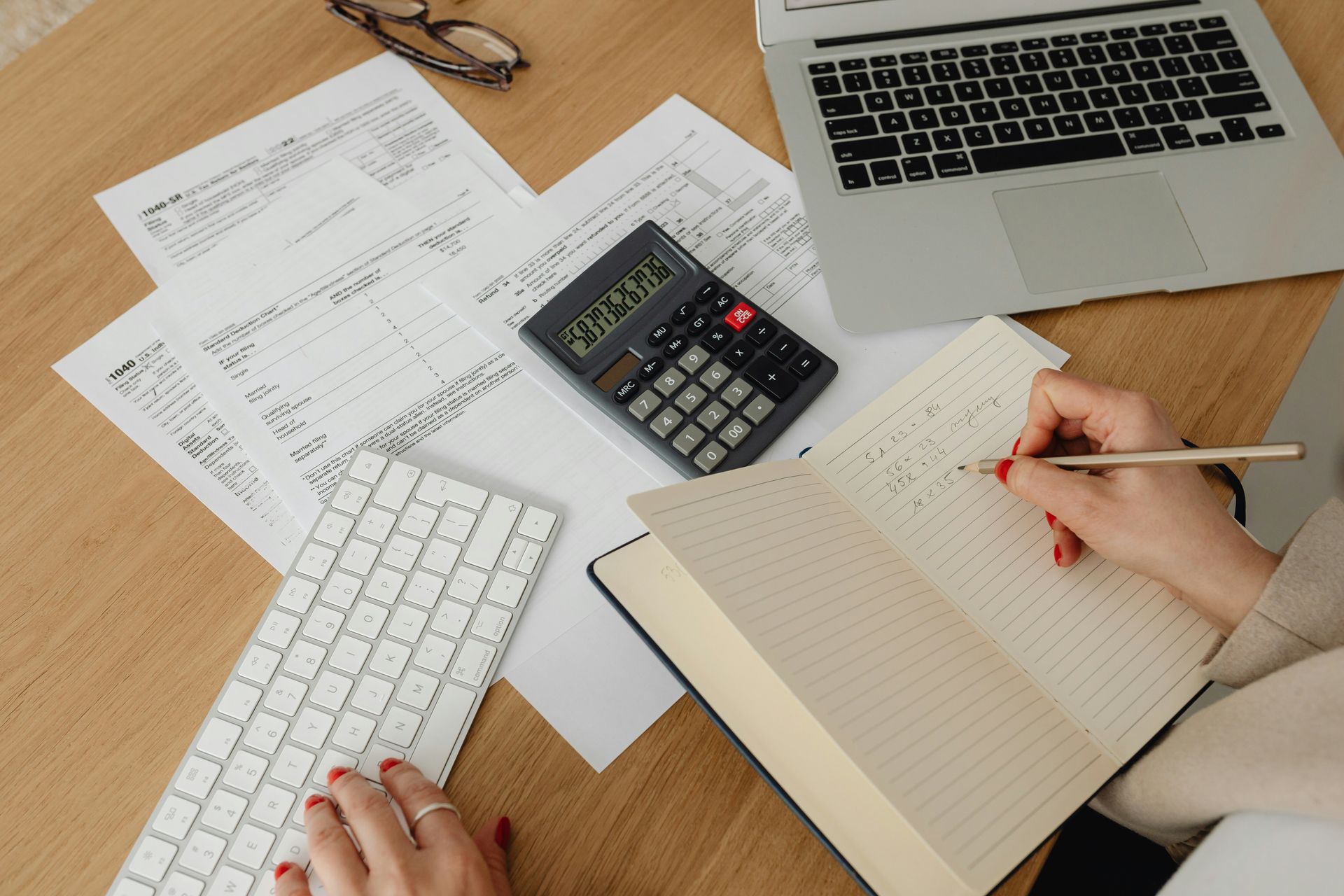 Hands working on taxes with a calculator, keyboard, laptop, papers, and notebook on a desk.