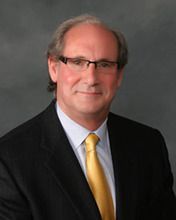 Man in glasses, suit, and gold tie, smiling against a dark gray backdrop.