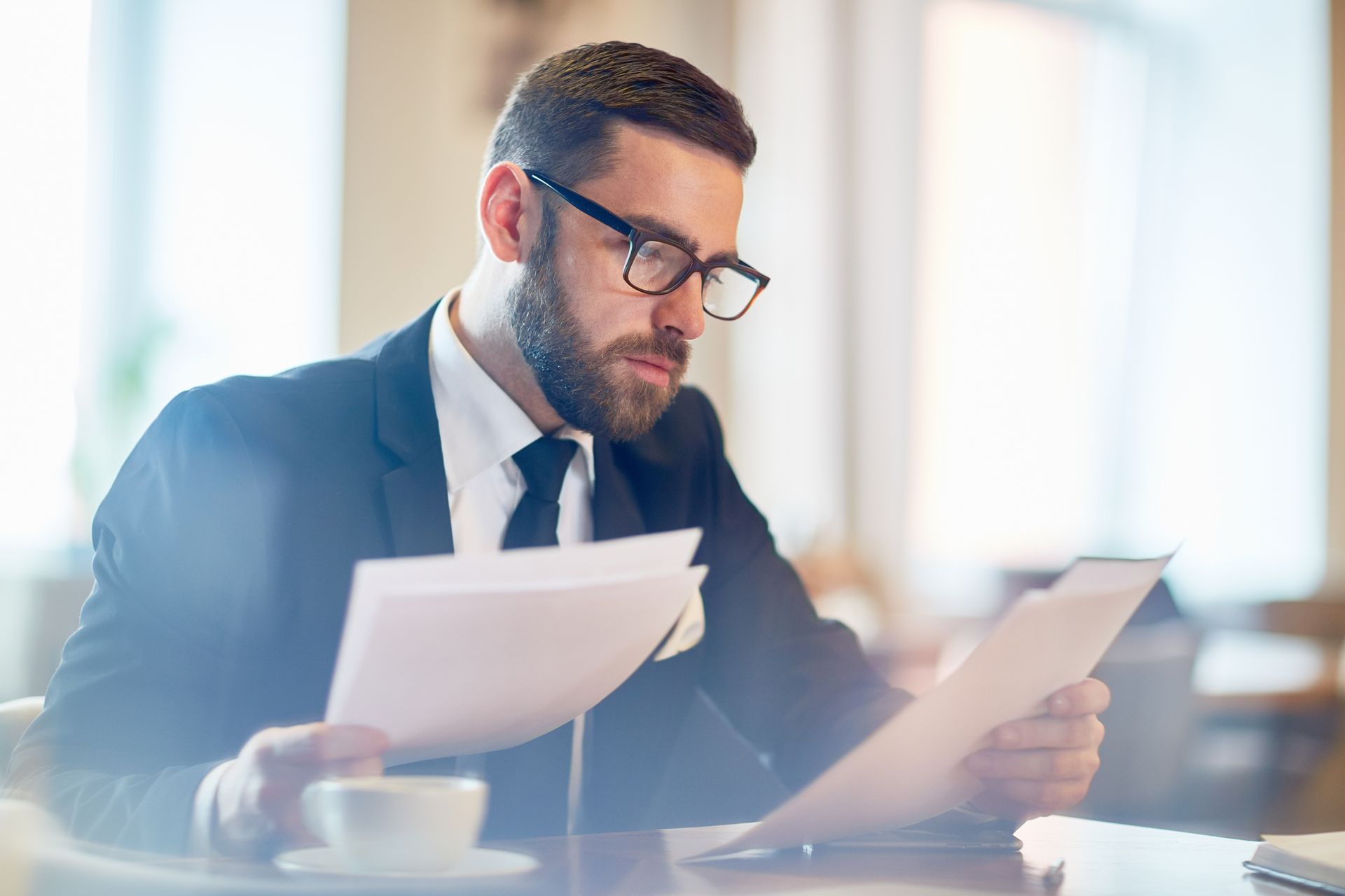 Man in suit and glasses reading papers at a table, coffee nearby.