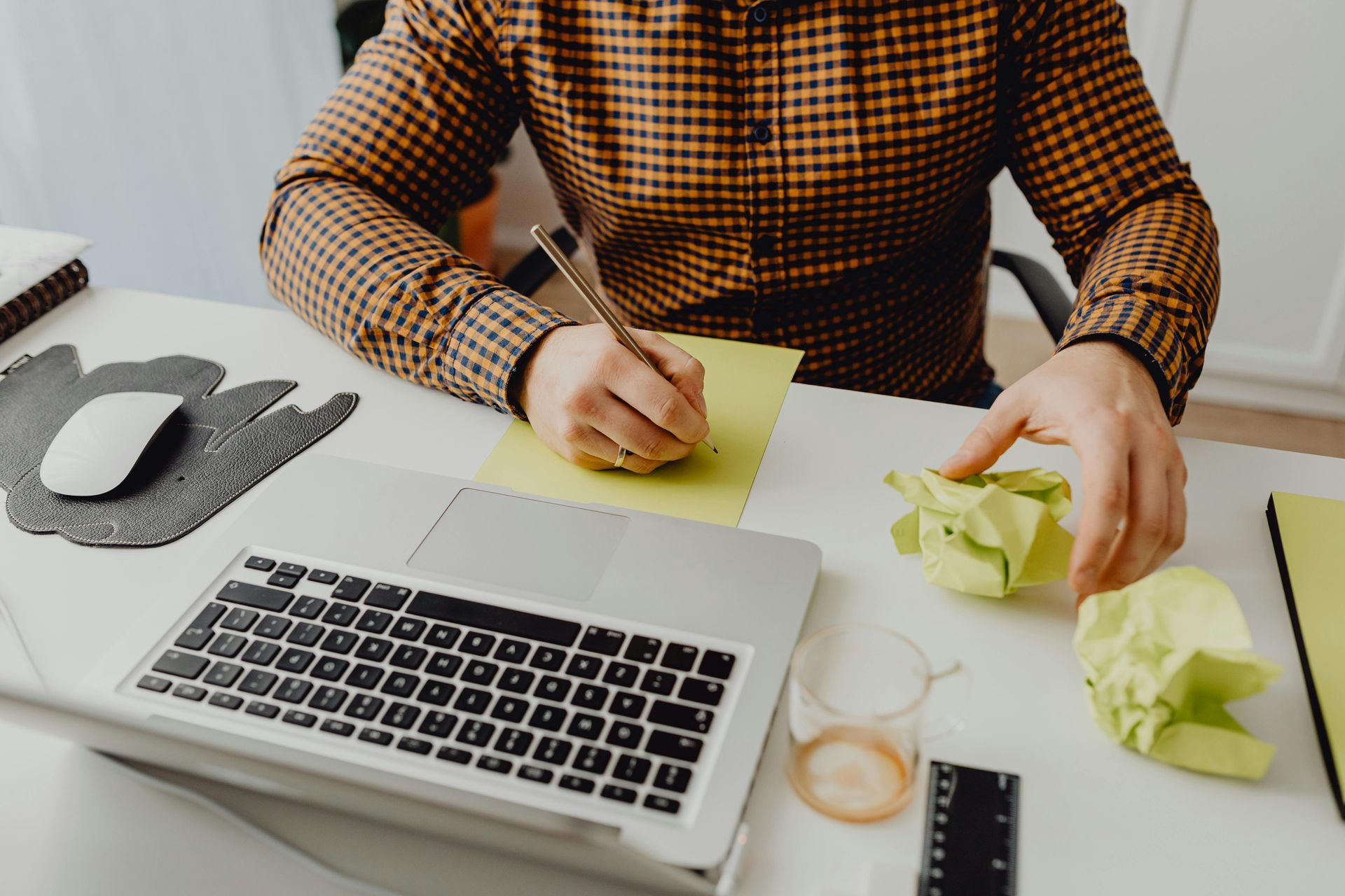 Person writing on yellow paper at a white desk with laptop, crumpled paper, and mouse.