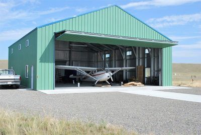 Green airplane hangar with an open door, housing a small airplane. A gravel driveway extends out.