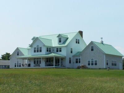 White farmhouse with green roof, porch, and dormers, set on a grassy field under a blue sky.
