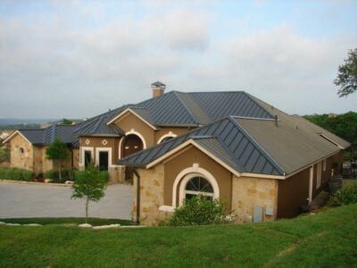 Tan stucco house with dark blue metal roof, stone accents, on a grassy hill.