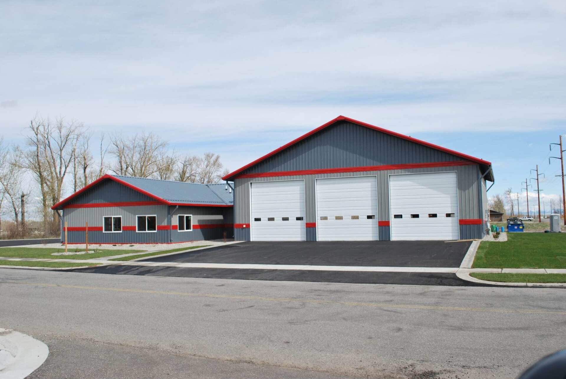 Gray and red building with three garage doors. Asphalt driveway. Green grass and blue sky.