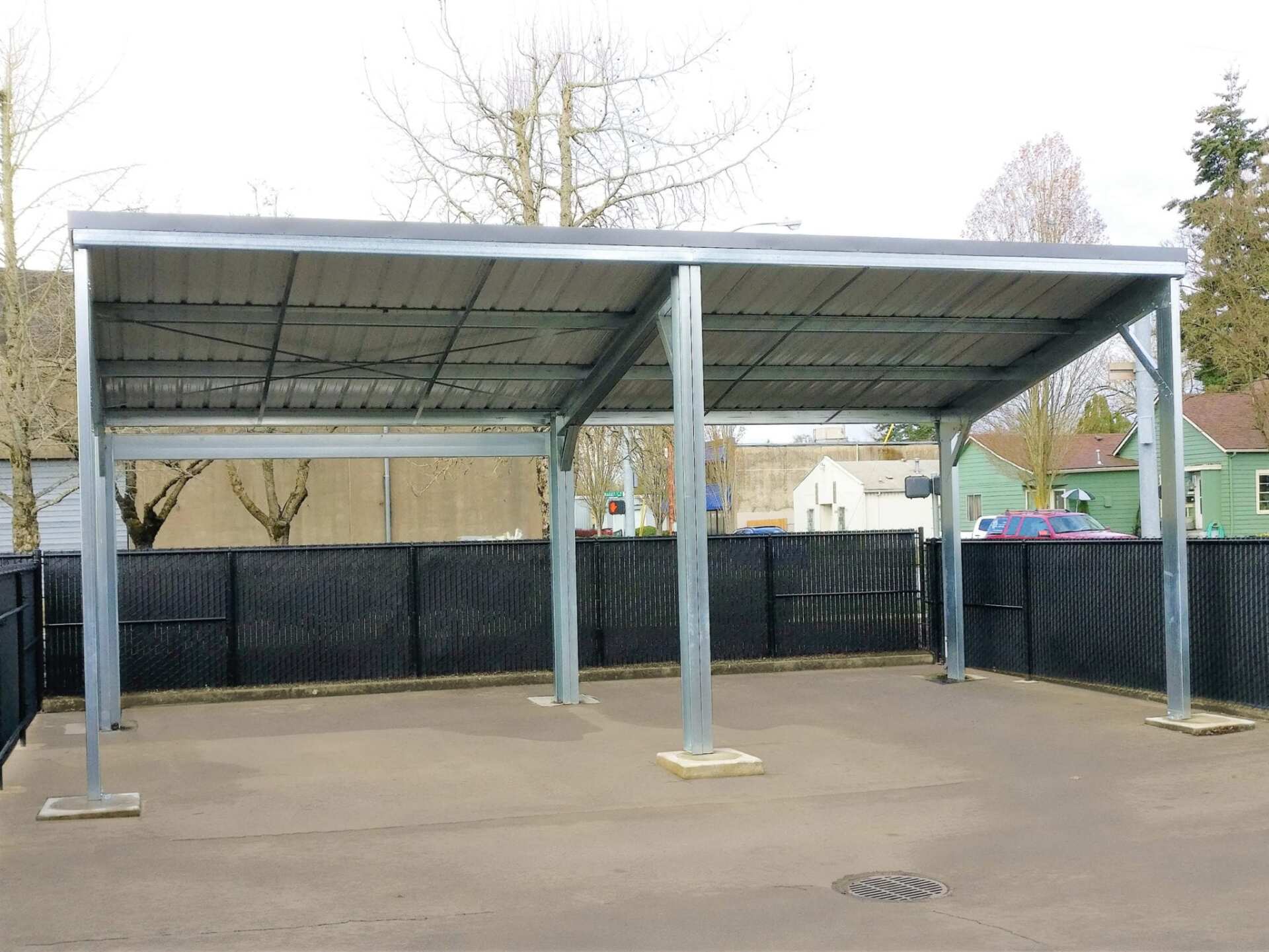 A metal carport with a corrugated metal roof, standing in a paved area.