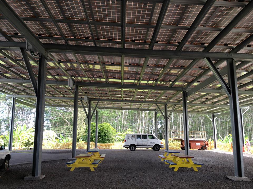Carport with solar panels overhead, picnic tables, and a van in a natural setting.