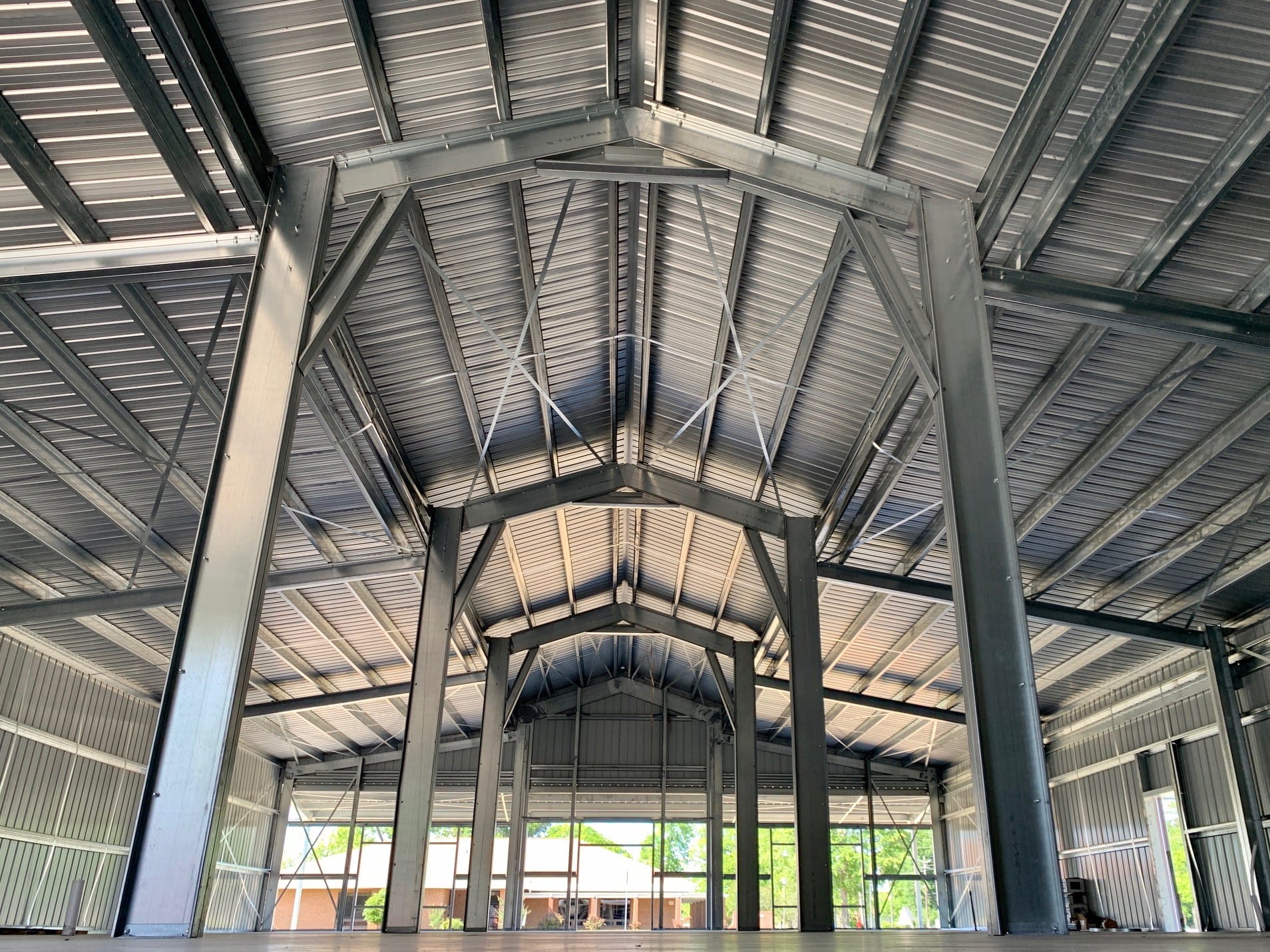 Interior view of a metal building under construction; metal beams, gray roof, windows, and a wooden floor.