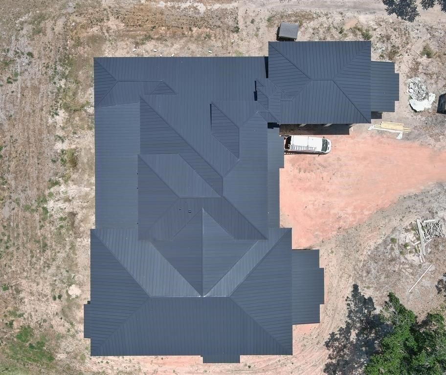 Overhead view of a dark gray metal roof on a house under construction; surrounded by dirt and some foliage.