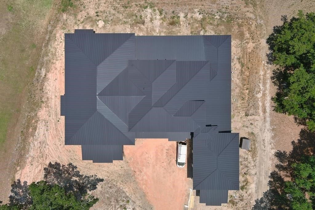 Overhead view of a dark roofed house and detached garage on a dirt lot, surrounded by trees.