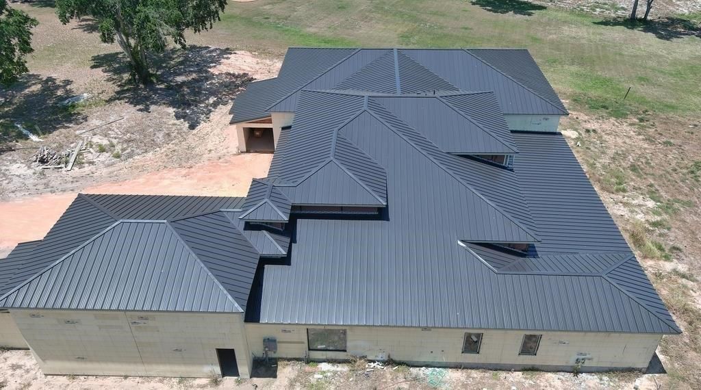 Overhead view of a gray-roofed house under construction in a dirt lot, surrounded by sparse trees.
