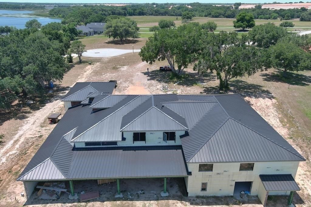 A large, light-colored house with a dark metal roof is under construction in a grassy field.