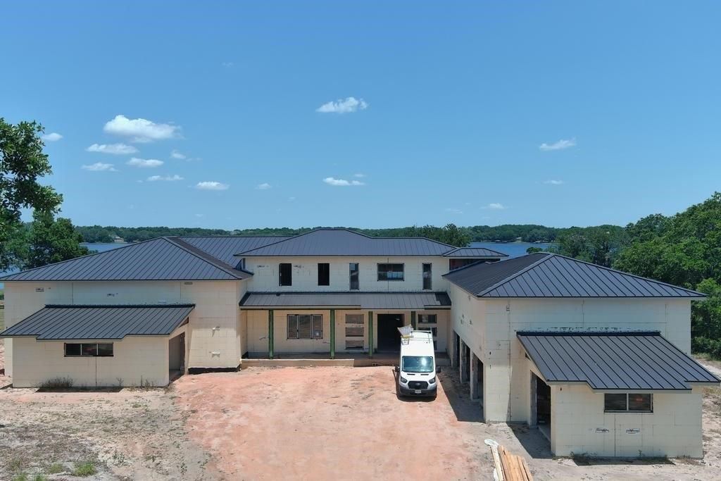 A partially built, beige multi-level house with a dark roof, white van parked out front, on a dirt lot with trees and water in the background.