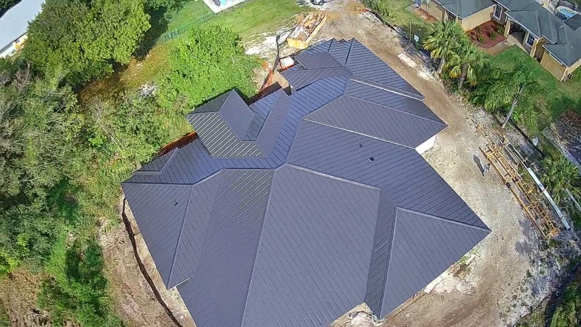 Aerial view of a dark gray metal roof of a house under construction, surrounded by dirt and vegetation.
