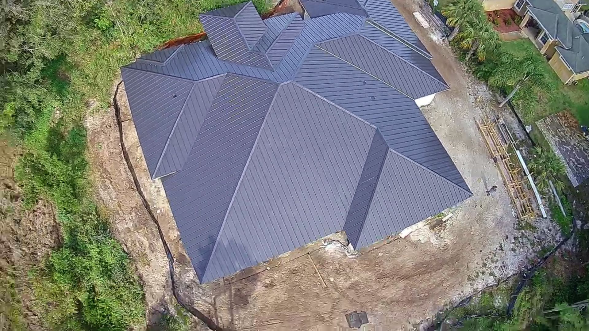Aerial view of a newly constructed house with a dark gray roof, surrounded by dirt and vegetation.
