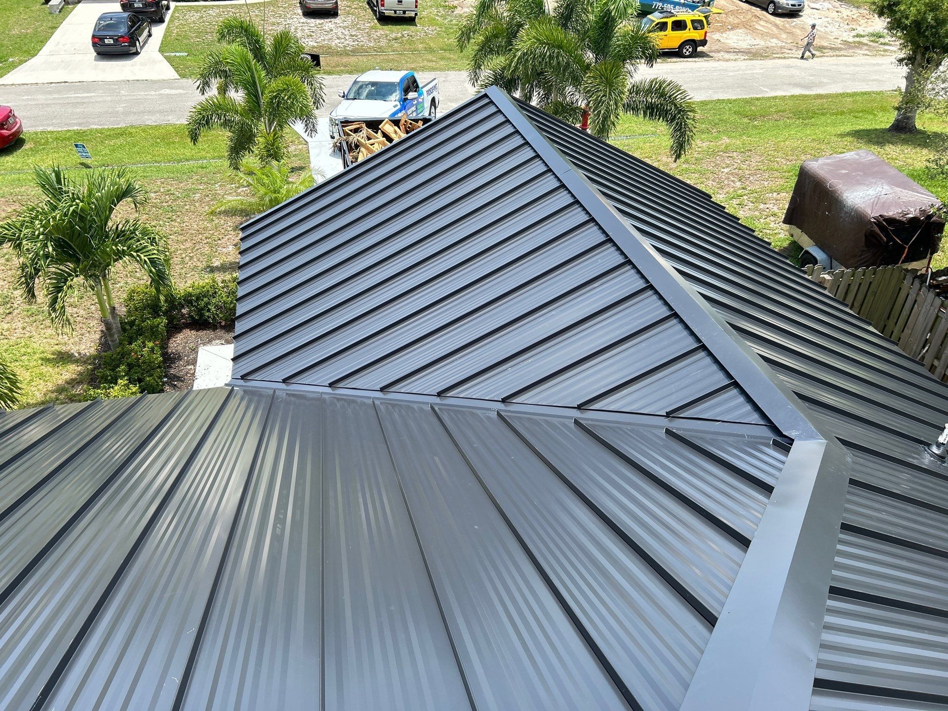 A metal roof with gray panels, viewed from above, with a partial view of the surrounding yard and street.