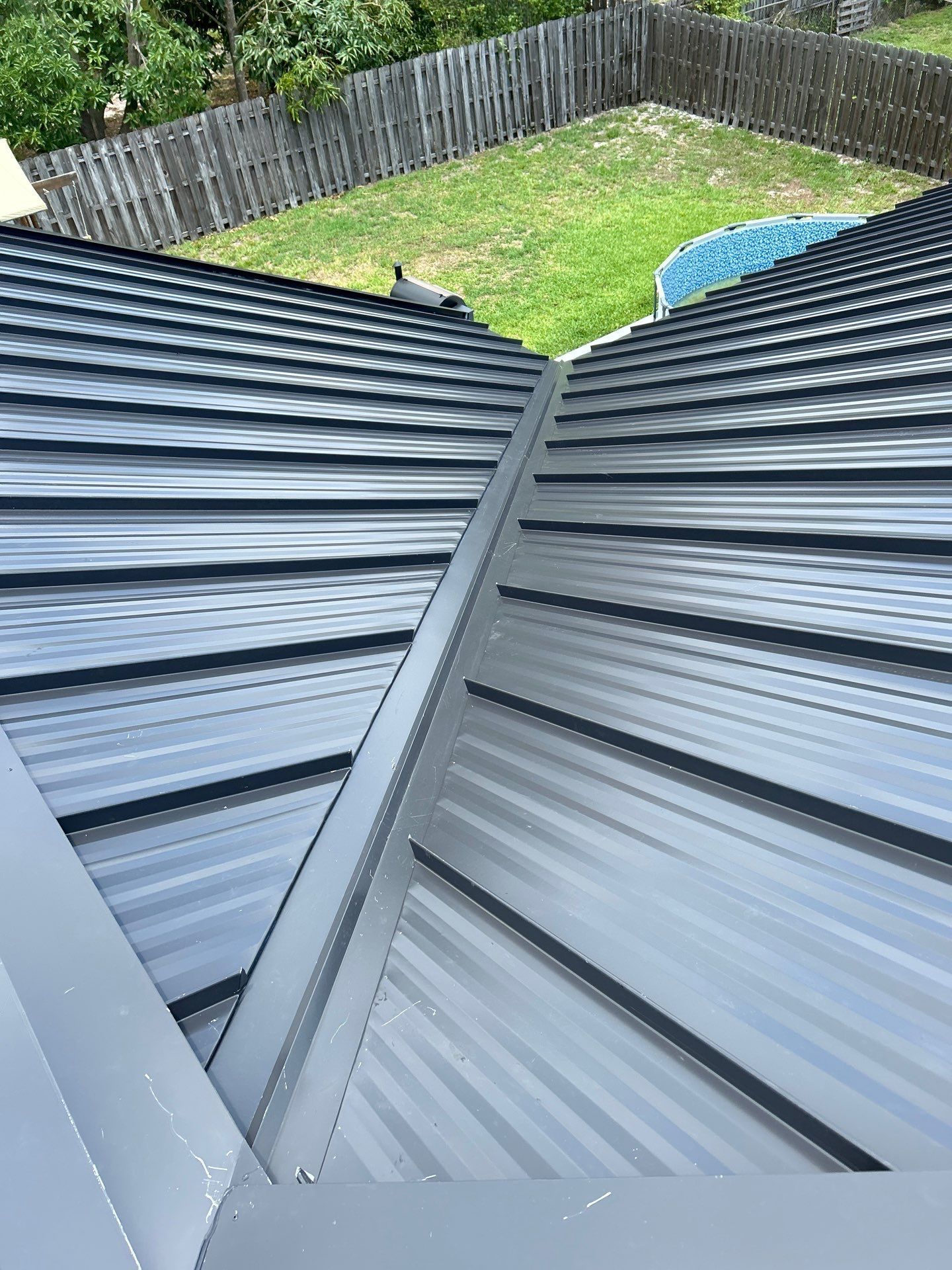 Close-up view of a dark metal roof with intersecting ridges. A green lawn and fence are visible in the background.