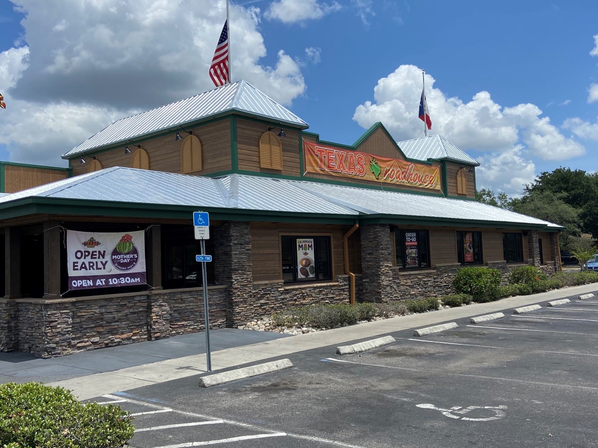 A restaurant with a stone facade and silver roof, US and Texas flags, parking lot, and blue handicap sign.