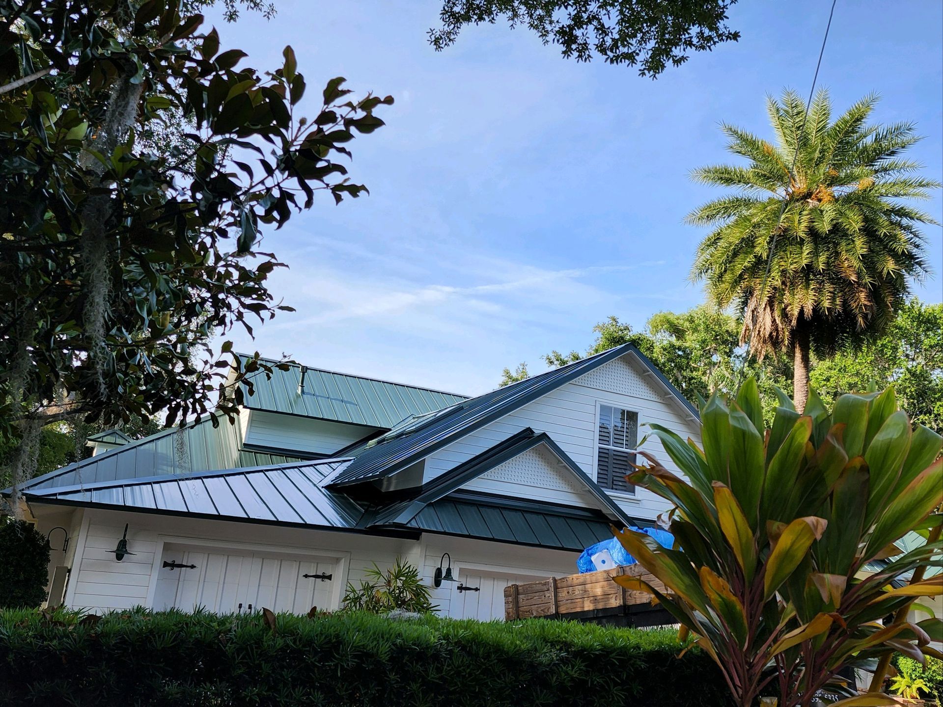 White house with green metal roof, blue sky, palm tree, and greenery.