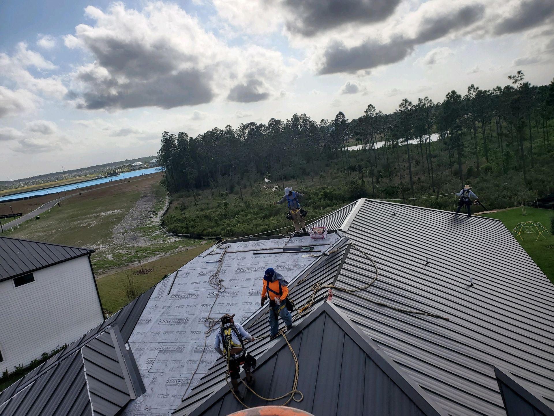 Roofers working on a gray metal roof, with harnesses and safety ropes. Cloudy sky, trees in the background.