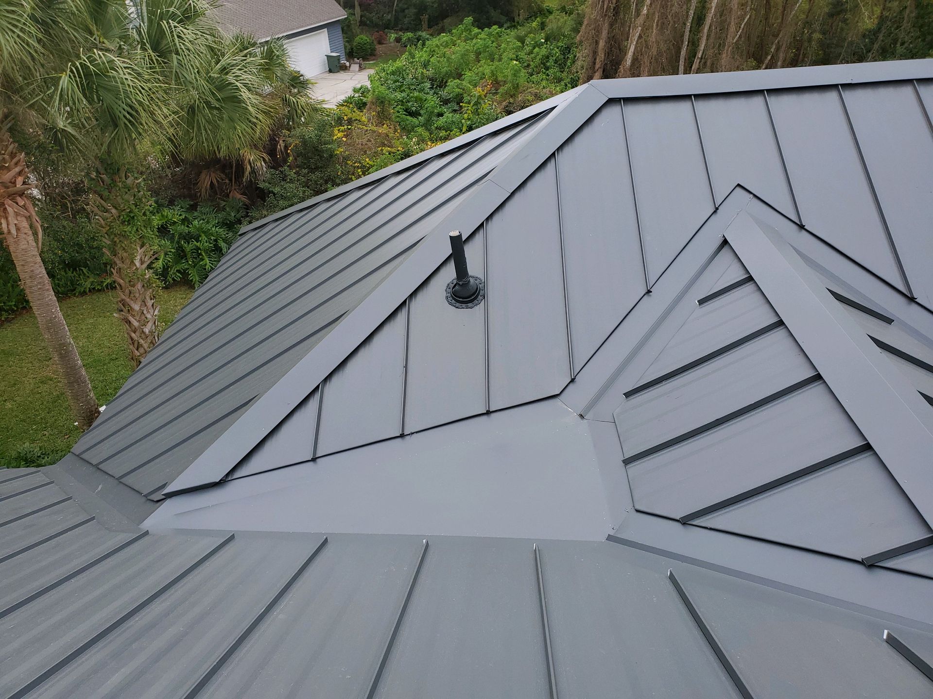 Gray metal roof with ridges, vent pipe, and dark trim, surrounded by greenery.