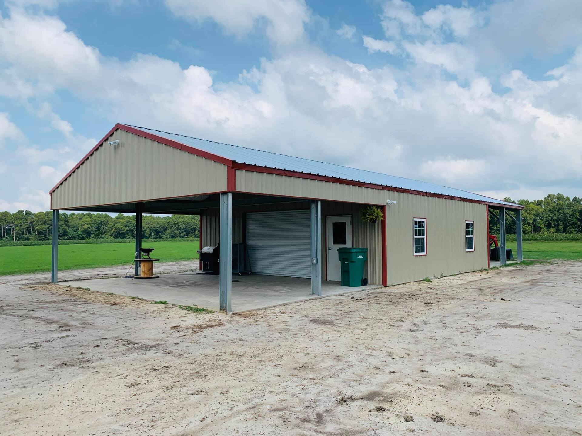 Beige metal building with a carport, red trim, and a white roll-up door in a field under a cloudy sky.