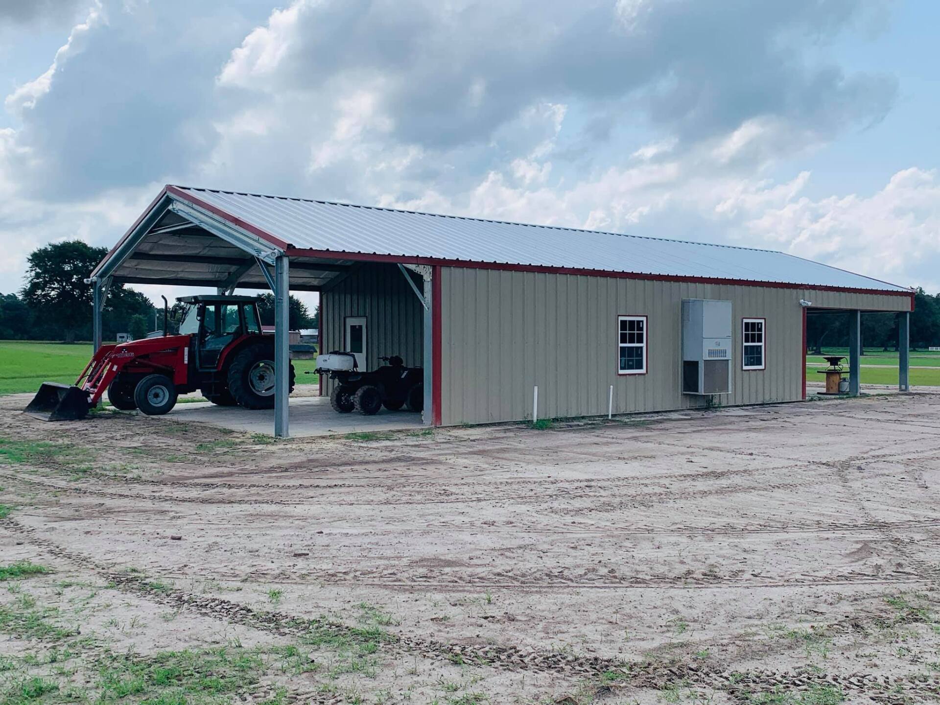 Beige metal barn with a red tractor parked beneath the roof, under a cloudy sky.