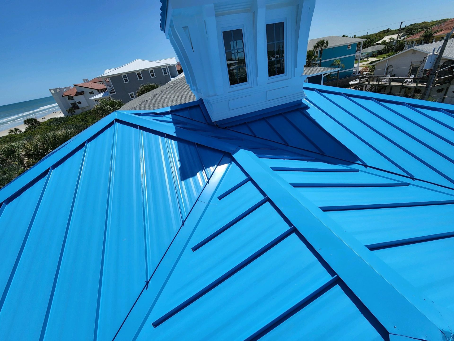Blue metal roof on a house with a white chimney against a sunny coastal backdrop.