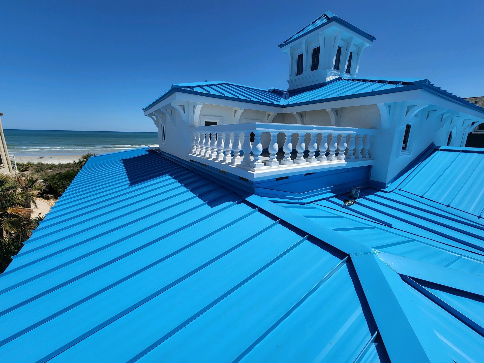 Blue metal roof on a white beach house with a balcony, overlooking the ocean on a sunny day.