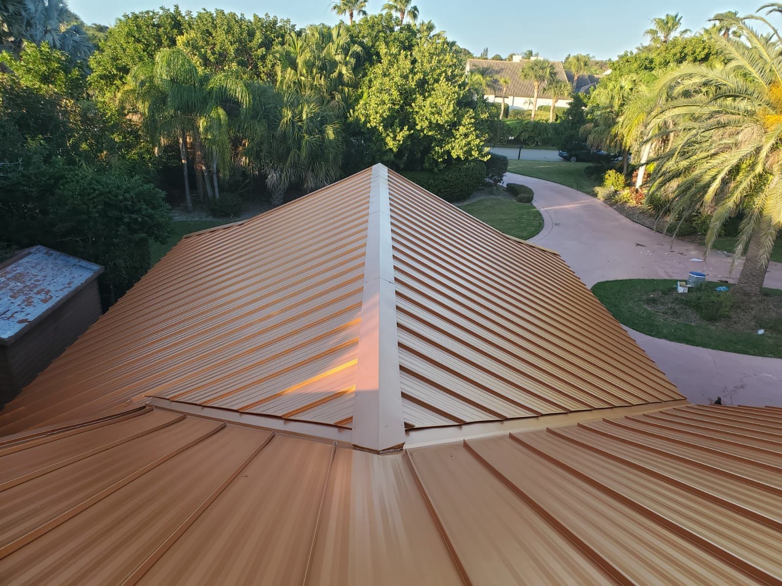 Bronze metal roof with prominent ridges, angled toward a central ridge. Trees and driveway in the background.