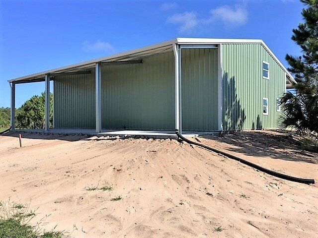 Green metal barn with a covered side, on a sandy area, under a blue sky.